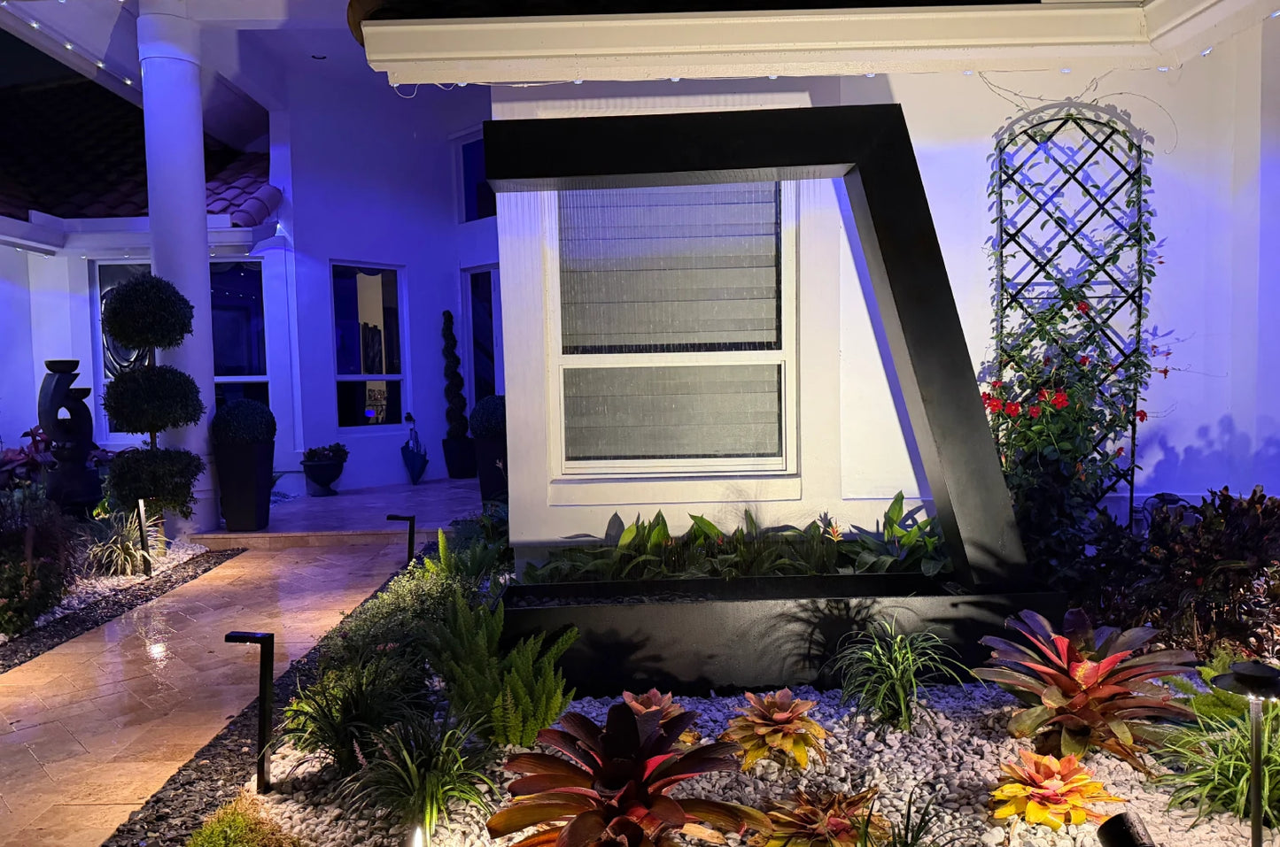 Illuminated black steel waterfall feature against a home's front wall at night, framed by bromeliads, white rock and a lit walkway