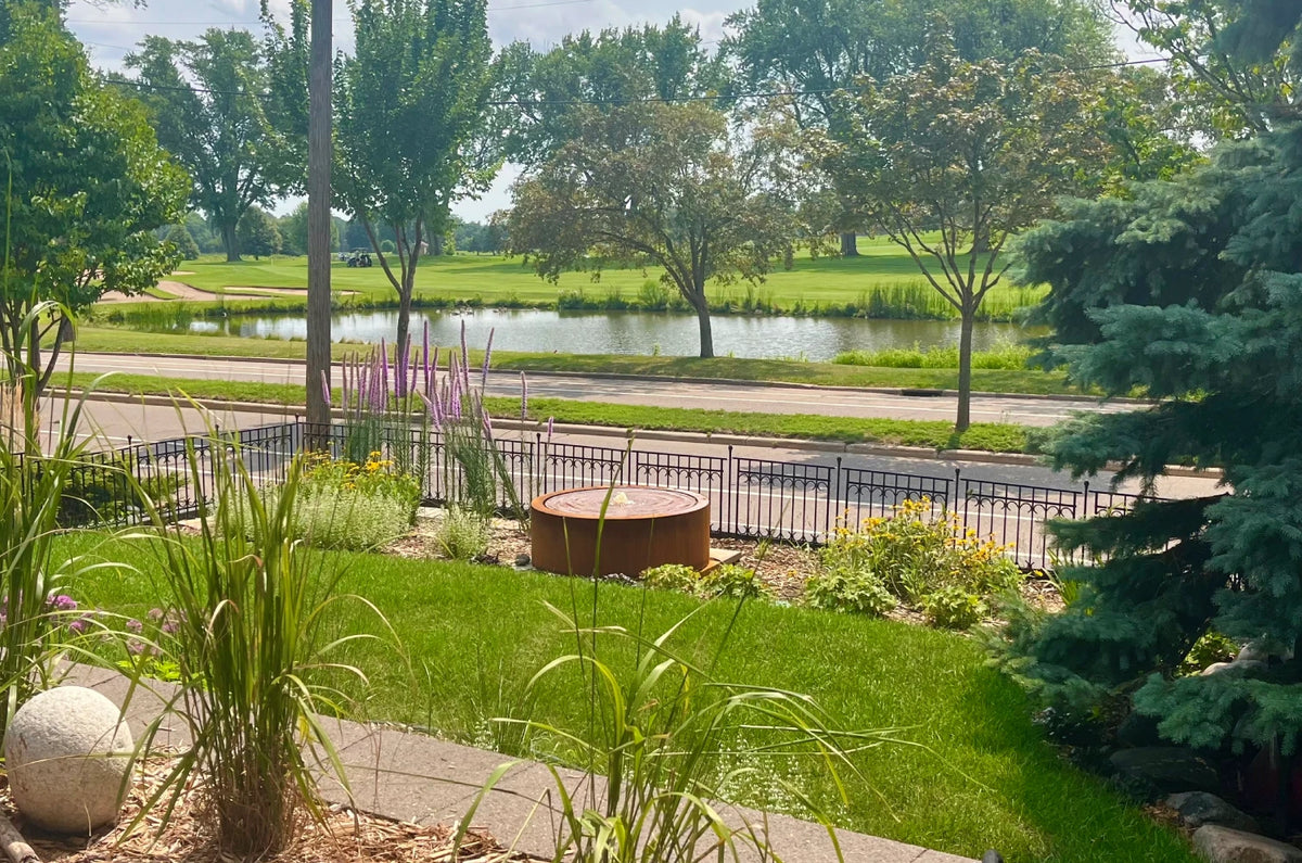 Round corten steel fountain in a front-yard perennial garden with wrought iron fence overlooking a golf course pond