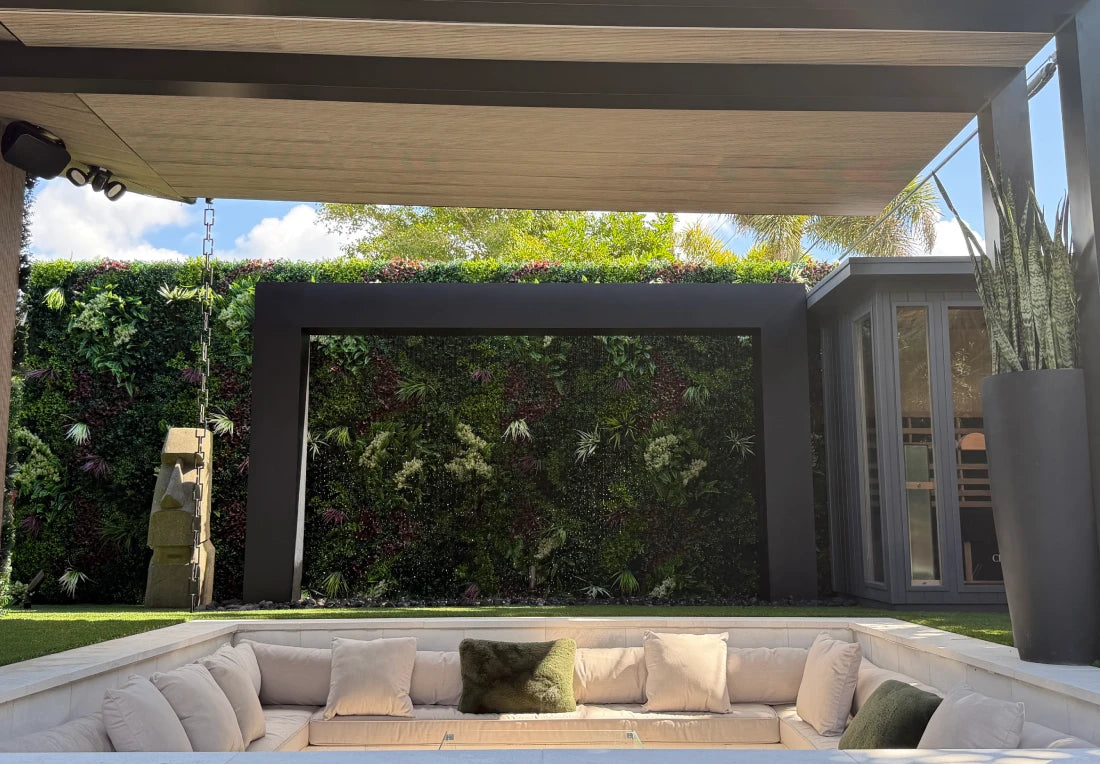 Black steel rain curtain fountain door built into a shaded pergola with a lush living plant wall backdrop above a sunken outdoor lounge seating area.