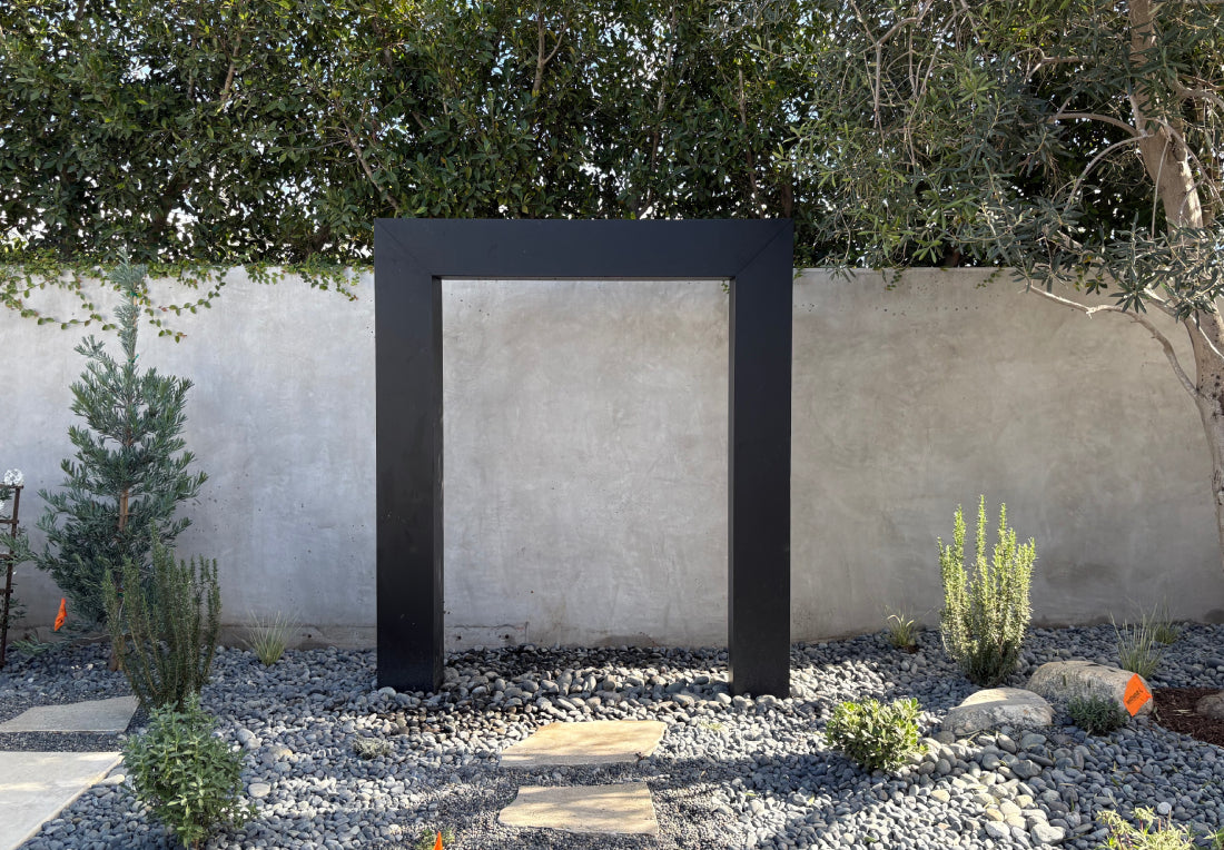Black powder-coated steel rain curtain fountain door installed against a stucco garden wall, surrounded by river rocks, native shrubs, and an olive tree, creating a refined modern outdoor water feature for garden and backyard design inspiration