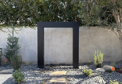 Black powder-coated steel rain curtain fountain door installed against a stucco garden wall, surrounded by river rocks, native shrubs, and an olive tree, creating a refined modern outdoor water feature for garden and backyard design inspiration