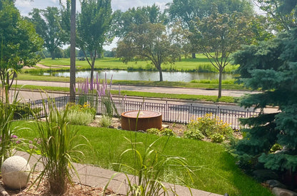 Round corten steel fountain in a front-yard perennial garden with wrought iron fence overlooking a golf course pond
