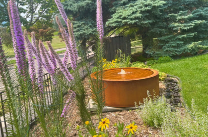 Rust-finished round steel fountain in a native pollinator garden with purple blazing star, black-eyed susans and blue spruce
