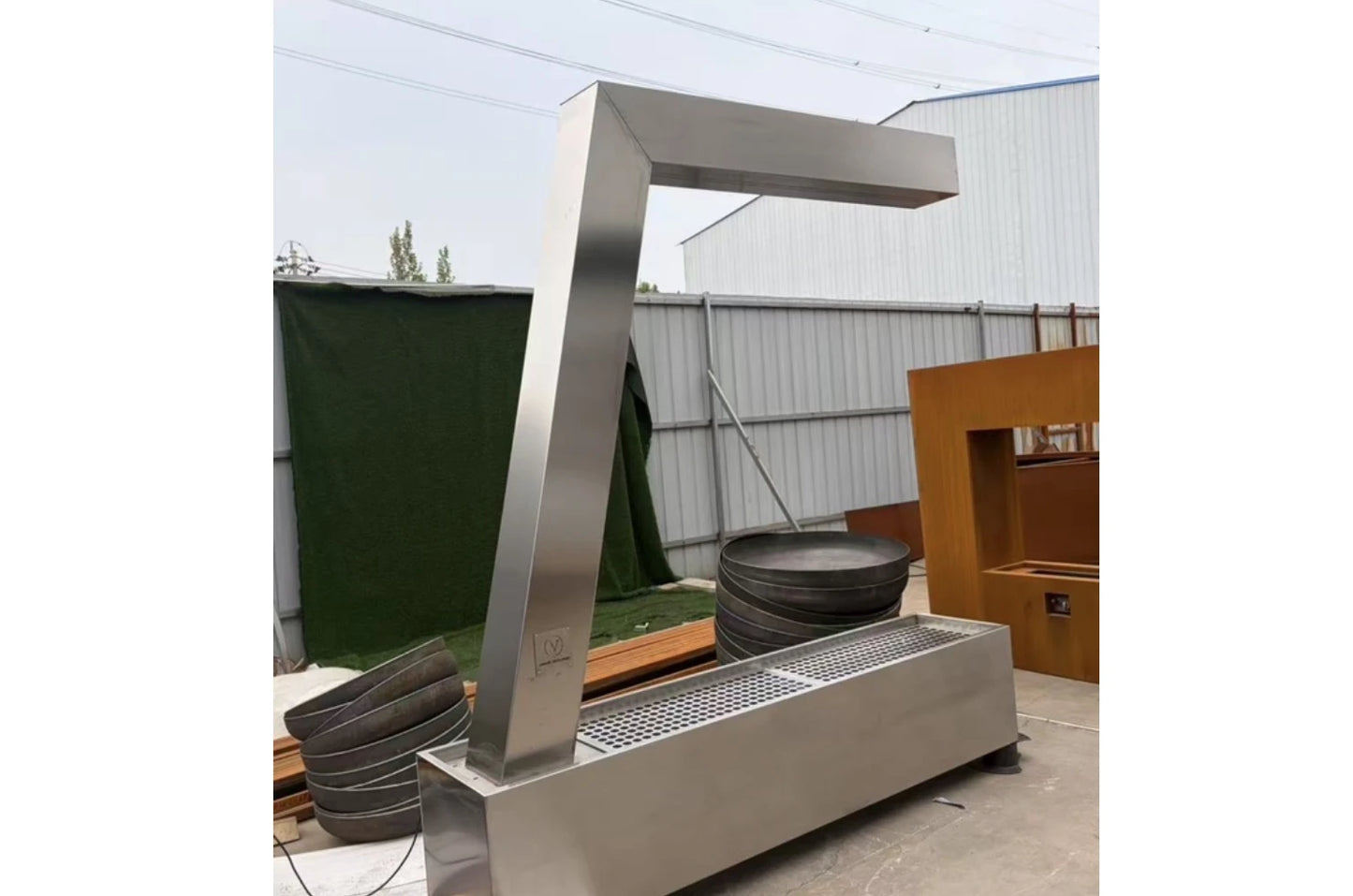 Brushed stainless steel L shaped waterfall frame with base grate in a fabrication yard, corten pieces and bowls behind