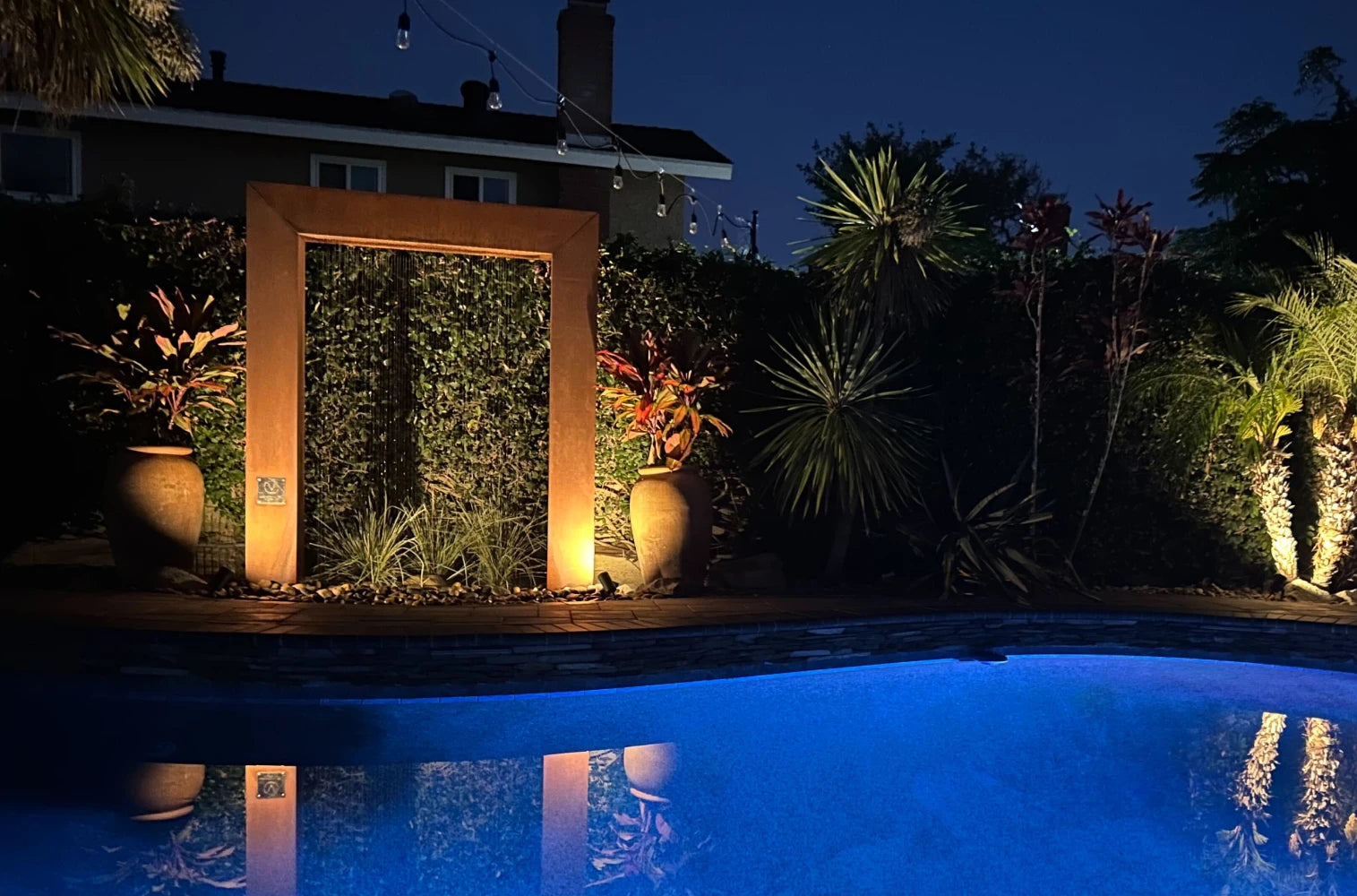 Illuminated rain curtain fountain door beside a glowing blue swimming pool at night, surrounded by tropical potted plants and string lights, creating an elegant poolside water feature and inspiring luxury backyard pool design ideas.
