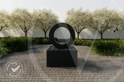 Modern matte black circular outdoor planter fountain with a ring water feature on a square pebble-topped base along a cobblestone garden path with flowering trees.