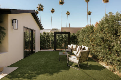 Black steel water feature frame on an artificial turf lawn beside a stucco home with patio seating and tall palms