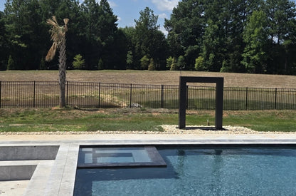 Black steel water feature standing across a travertine pool deck with an in-ground pool, spa and wooded backdrop