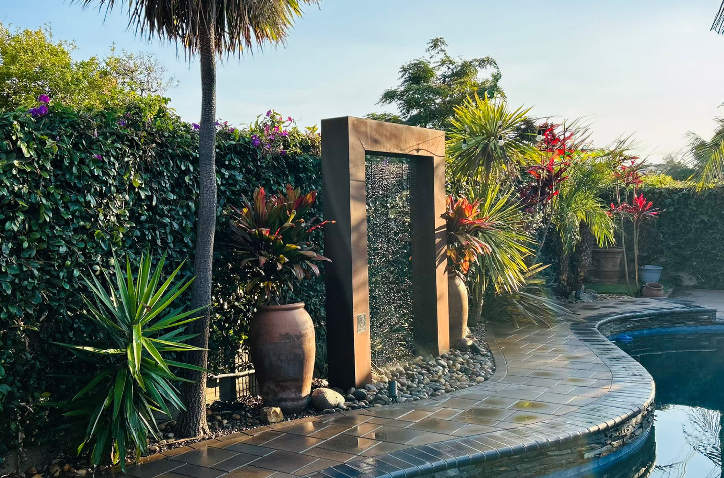 Rain curtain fountain door flanked by large terracotta planters and palm trees along a curved stone walkway beside a backyard swimming pool.