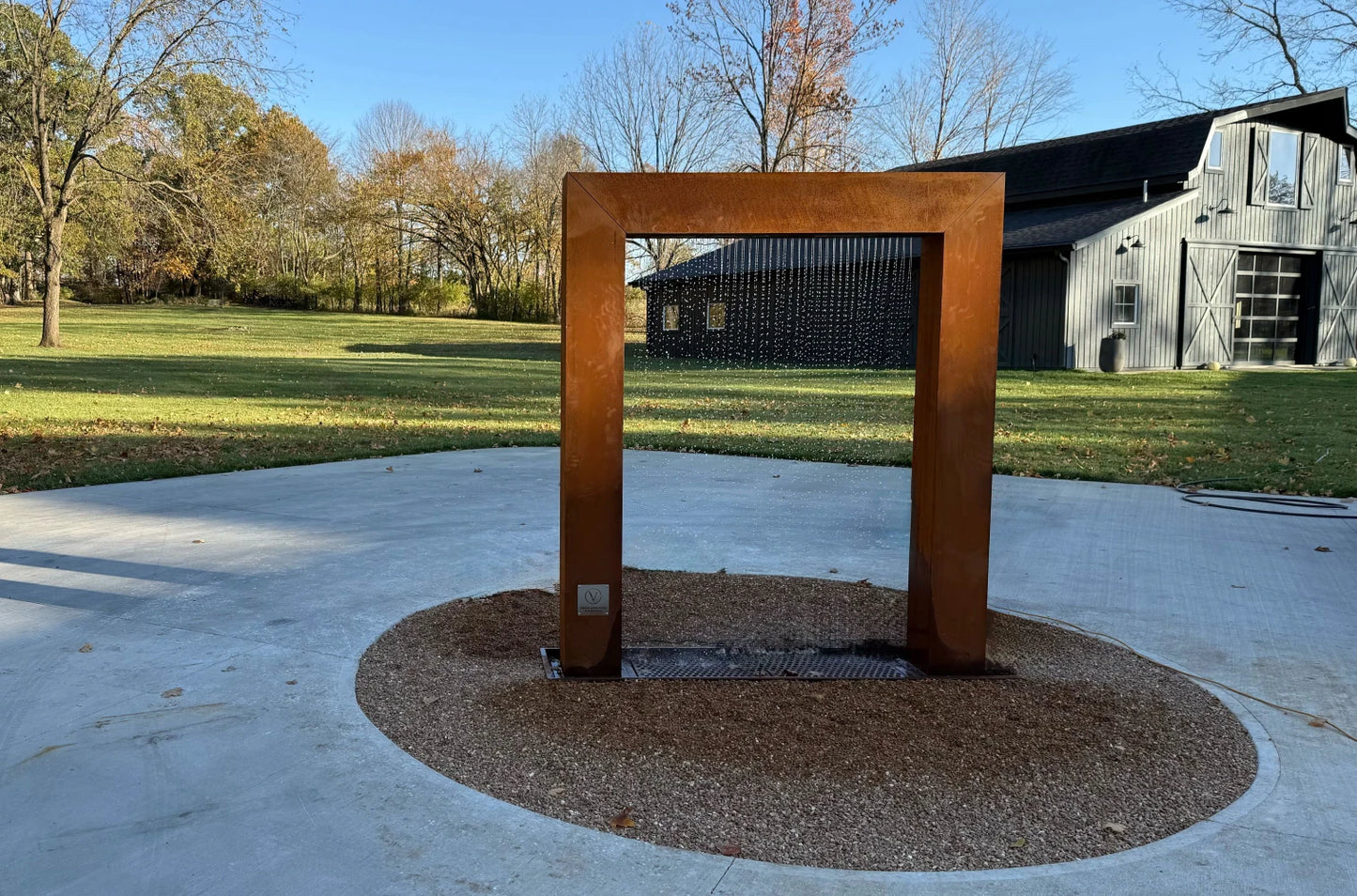 Corten steel rain curtain fountain door installed on a concrete patio with a decorative gravel border, featuring flowing water in bright daylight beside a rustic barn-style home and creating a distinctive modern farmhouse backyard water feature.