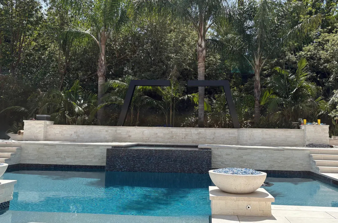 Two Valyrian Designs L Model black powder-coated steel rain curtain fountain frames rising above a tiled pool terrace wall, surrounded by tall palm trees and a modern fire bowl on a sunny day, creating a striking luxury backyard pool and outdoor living design.