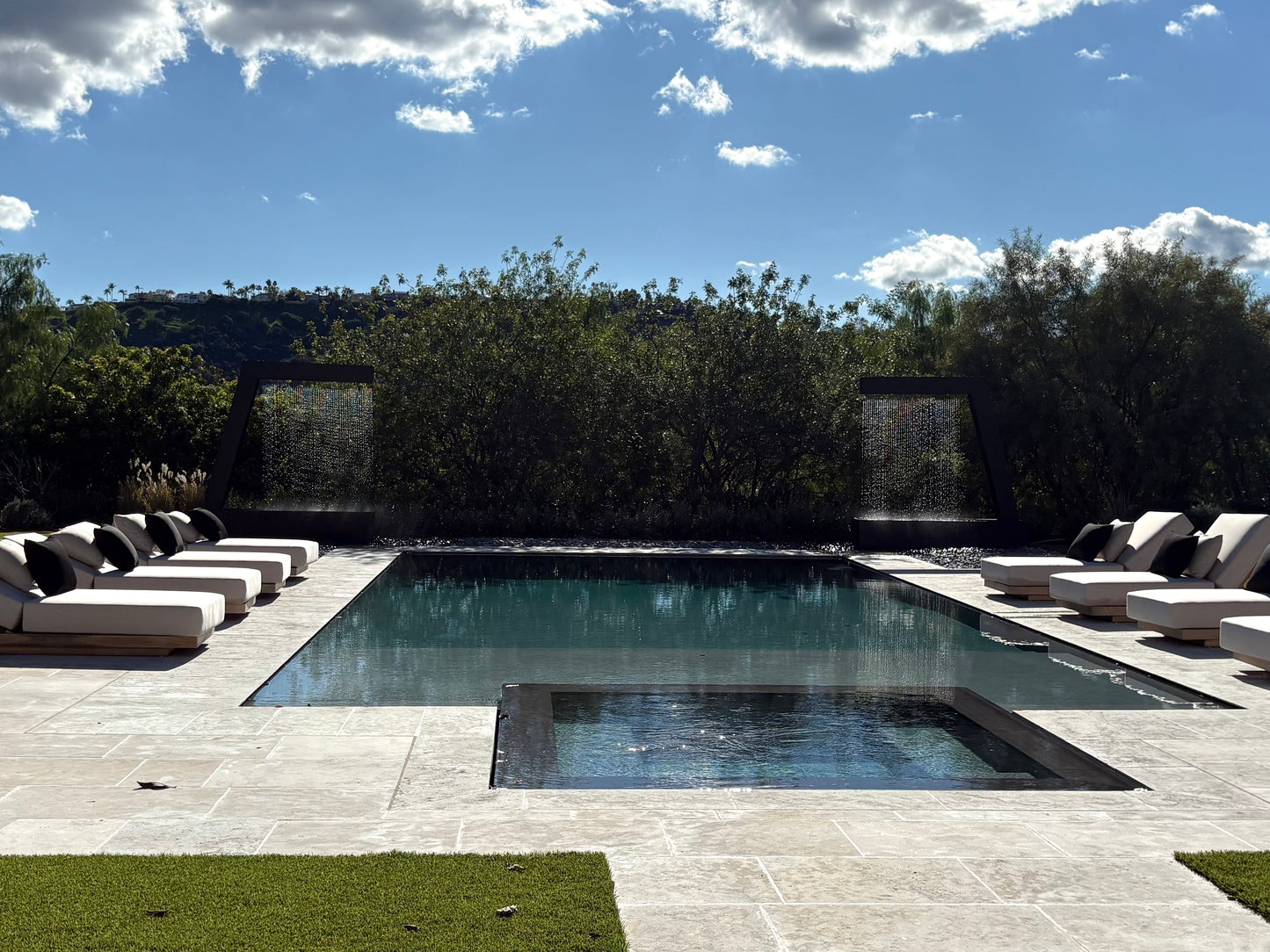 Resort-style luxury backyard pool lined with white lounge chairs and framed by two Valyrian Designs L Model black powder-coated steel rain curtain fountains, cascading against a hillside tree line beneath a clear blue sky for inspiring poolside landscape and outdoor living ideas