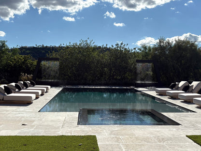 Resort-style luxury backyard pool lined with white lounge chairs and framed by two Valyrian Designs L Model black powder-coated steel rain curtain fountains, cascading against a hillside tree line beneath a clear blue sky for inspiring poolside landscape and outdoor living ideas
